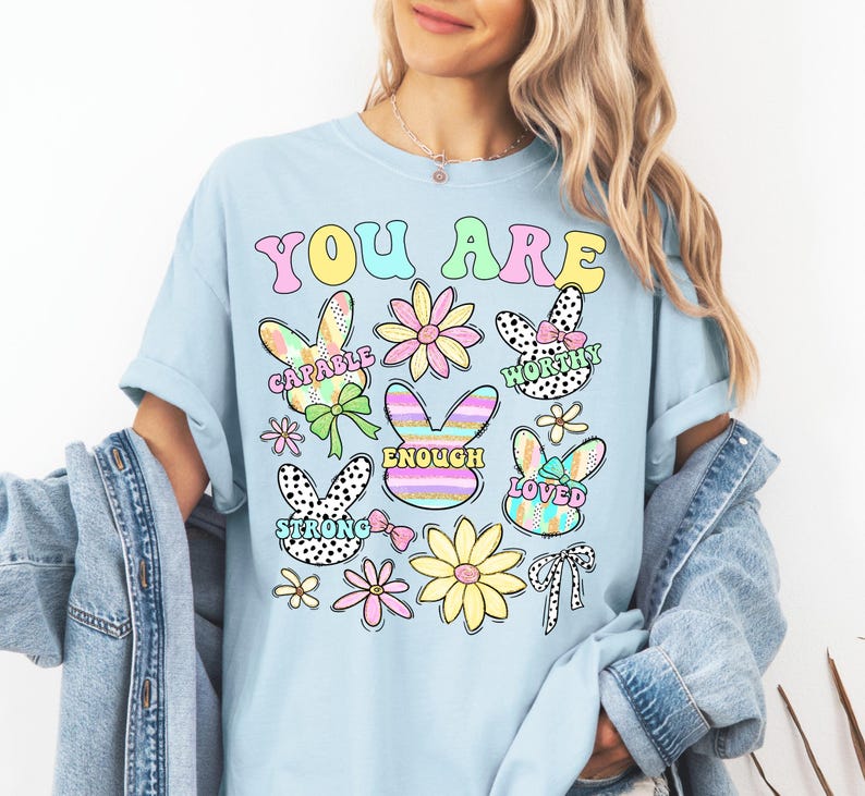 May include: Light blue t-shirt with the text "You Are" in rainbow colors. The shirt features a variety of colorful bunny and flower illustrations with positive affirmations like "Capable", "Worthy", "Enough", "Loved", and "Strong".