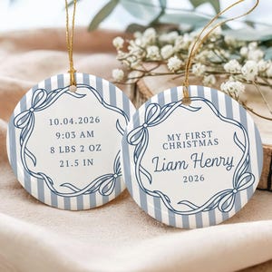May include: Two round ceramic ornaments with a light blue and white vertical striped pattern. Each features a dark blue decorative ribbon border. One displays birth details: 10.04.2026, 9:03 AM, 8 lbs 2 oz, 21.5 in. The other reads My First Christmas, Liam Henry, 2026.
