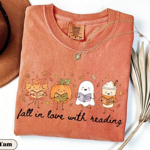 Peut inclure: Un t-shirt &agrave; col rond couleur rouille avec un motif repr&eacute;sentant une feuille d'&eacute;rable, une citrouille, un fant&ocirc;me et une tasse de caf&eacute;, chacun tenant un livre. Le texte indique fall in love with reading. &Eacute;tiquette Comfort Colors au col.