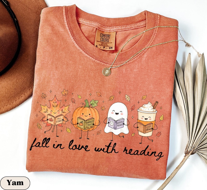 Peut inclure: Un t-shirt &agrave; col rond couleur rouille avec un motif repr&eacute;sentant une feuille d'&eacute;rable, une citrouille, un fant&ocirc;me et une tasse de caf&eacute;, chacun tenant un livre. Le texte indique fall in love with reading. &Eacute;tiquette Comfort Colors au col.