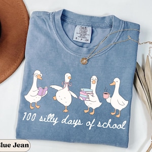 May include: Blue jean coloured t-shirt featuring a graphic of four cartoon geese. The geese are in different poses, holding books and a coffee cup. The text "100 silly days of school" is printed below the geese.