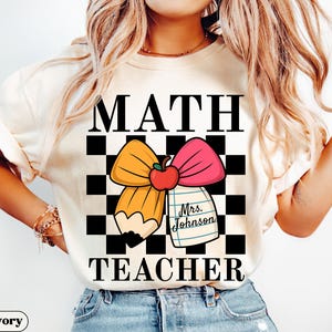 May include: Ivory-colored t-shirt with the words "MATH TEACHER" in black block letters. A pencil and pink bow design with an apple and a notepad that says "Mrs. Johnson" is in the center. The background is a black and white checkered pattern.