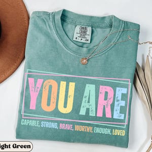 May include: A light green t-shirt with the words "YOU ARE" in a colorful, distressed font. Below, the shirt reads "CAPABLE, STRONG, BRAVE, WORTHY, ENOUGH, LOVED." A silver necklace with a pendant rests on the shirt. The shirt is folded.