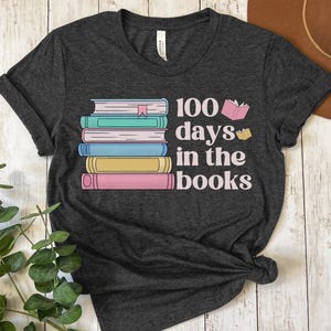 Pu&ograve; includere: Una maglietta grigia scura con una pila di libri colorati e il testo "100 giorni tra i libri".