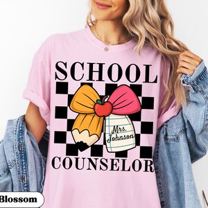 May include: Pink t-shirt with the words "SCHOOL COUNSELOR" in black block letters. The design includes a pencil and pink bow with an apple, set against a black and white checkered background. A tag reads "Mrs. Johnson".