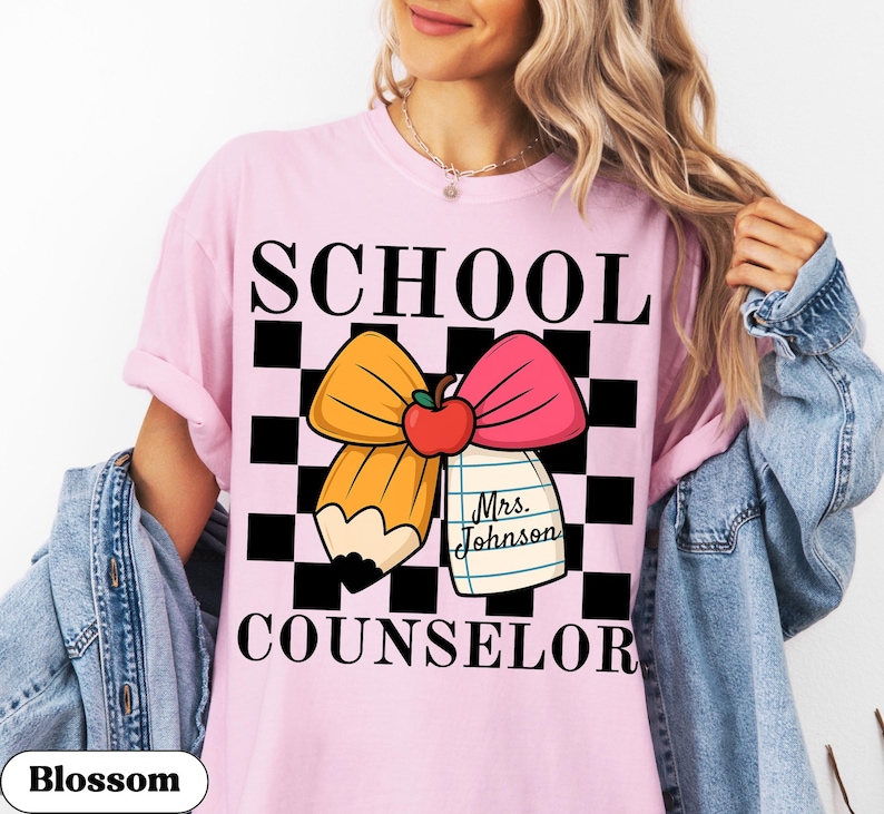 May include: Pink t-shirt with the words "SCHOOL COUNSELOR" in black block letters. The design includes a pencil and pink bow with an apple, set against a black and white checkered background. A tag reads "Mrs. Johnson".