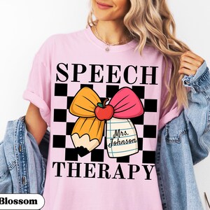 May include: Pink t-shirt with the words "SPEECH THERAPY" in black block letters. A cartoon pencil and pink bow with an apple are in the center. A small notepad with the name "Mrs. Johnson" is attached to the bow. The shirt has a black and white checkered background.
