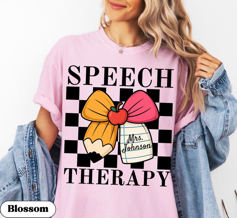 May include: Pink t-shirt with the words "SPEECH THERAPY" in black block letters. A cartoon pencil and pink bow with an apple are in the center. A small notepad with the name "Mrs. Johnson" is attached to the bow. The shirt has a black and white checkered background.
