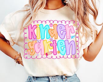 Puede incluir: Camiseta color crema con un gráfico colorido de "Kindergarten". El texto está en letras de burbujas con una variedad de colores, incluyendo rosa, azul, naranja y morado. El diseño incluye ilustraciones escolares como tijeras, un lápiz y un avión de papel.