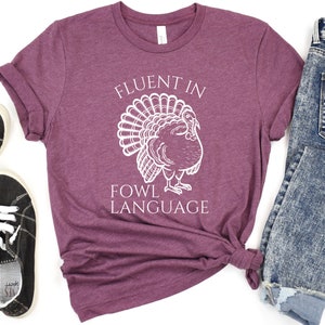 May include: A maroon t-shirt featuring the text "FLUENT IN FOWL LANGUAGE" and a white turkey illustration. The shirt is styled with blue jeans and black trainers.