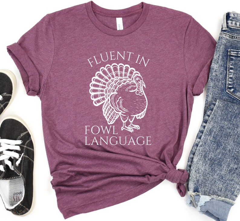 May include: A maroon t-shirt featuring the text "FLUENT IN FOWL LANGUAGE" and a white turkey illustration. The shirt is styled with blue jeans and black trainers.