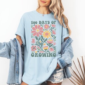 May include: A light blue t-shirt with a graphic of colorful flowers and the text "100 Days of Growing" in a retro font.
