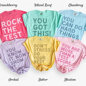 May include: Six colorful t-shirts in shades of pink, teal, blue, purple, yellow, and pink. Each shirt has a different motivational phrase printed in a contrasting color. The shirts are labeled with their color names.