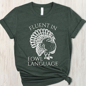 May include: Dark green t-shirt featuring the text "FLUENT IN FOWL LANGUAGE" and a white turkey illustration. The shirt has rolled sleeves and a knot at the bottom.