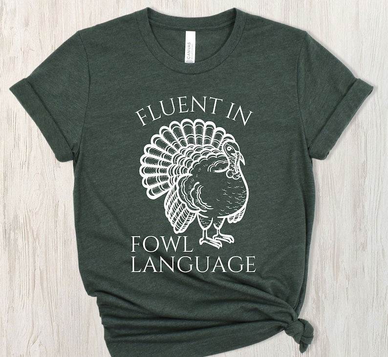 May include: Dark green t-shirt featuring the text "FLUENT IN FOWL LANGUAGE" and a white turkey illustration. The shirt has rolled sleeves and a knot at the bottom.