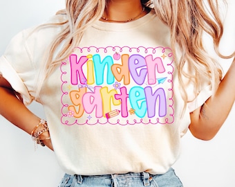 Puede incluir: Camiseta color crema con un gráfico colorido de "Kindergarten". El gráfico presenta letras de burbujas en varios colores, rodeadas por un borde rosa con elementos decorativos como tijeras, un avión de papel y un lápiz.