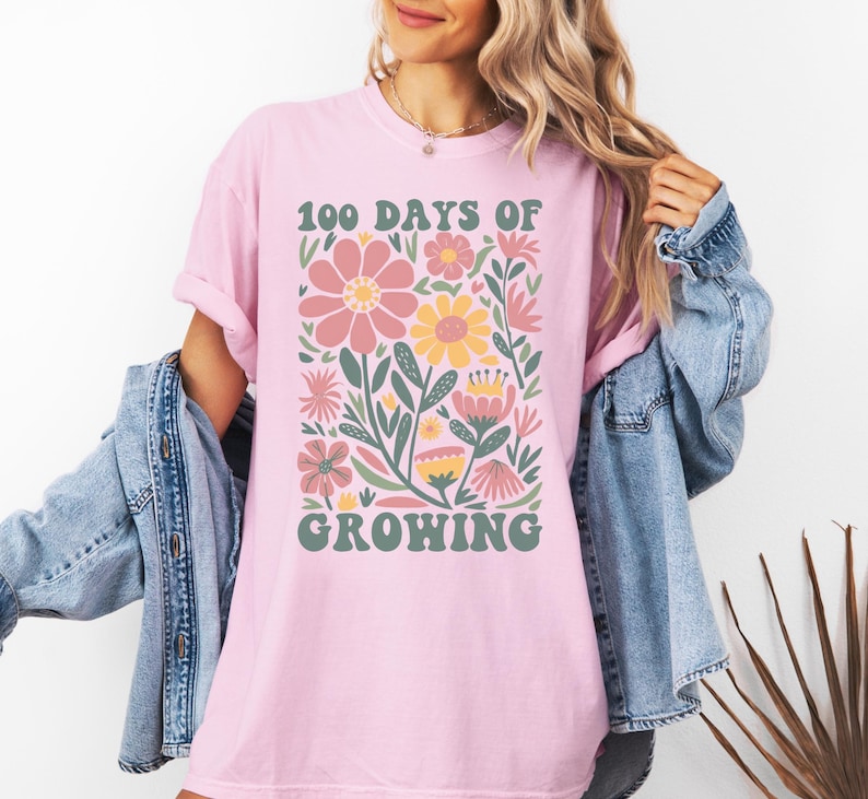 May include: Pink t-shirt with a floral design and the text "100 Days of Growing" in a retro font.