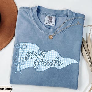 May include: A blue jean colored t-shirt featuring a light blue and white gingham patterned pennant flag graphic with two bows. The text "first grade" is written in a white script font across the flag. A gold necklace rests on the shirt.