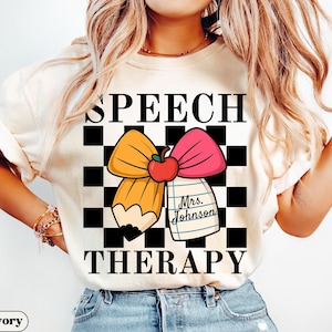 May include: Ivory-colored t-shirt with the words "SPEECH THERAPY" in black block letters. The design includes a pencil, a pink bow, and a red apple on a black and white checkered background. A notepad with the name "Mrs. Johnson" is also included.