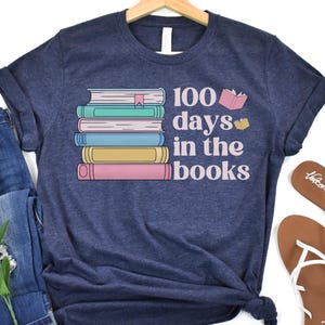 Pu&ograve; includere: Una maglietta blu navy con una pila di libri colorati e il testo "100 giorni tra i libri".