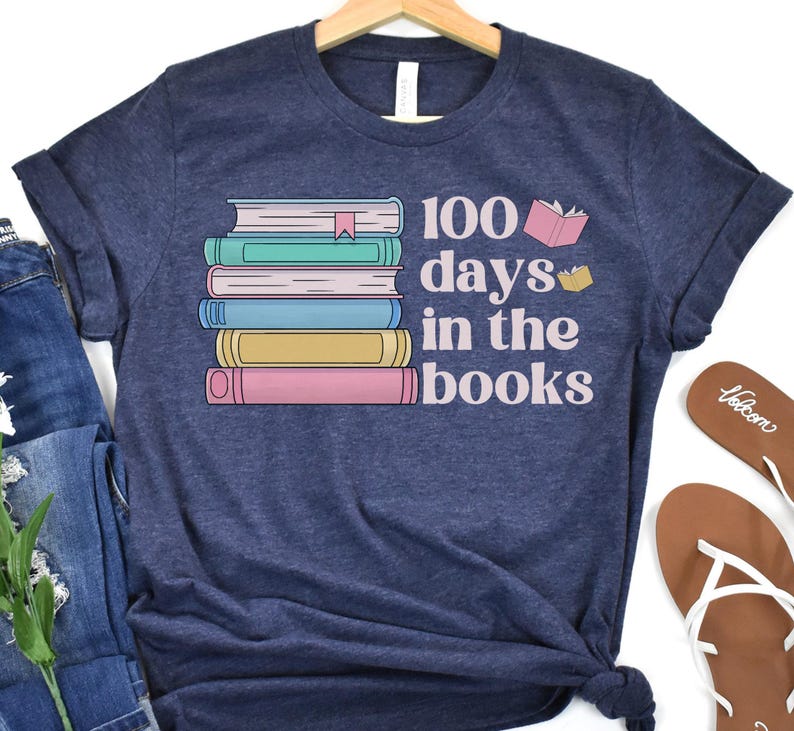 Pu&ograve; includere: Una maglietta blu navy con una pila di libri colorati e il testo "100 giorni tra i libri".