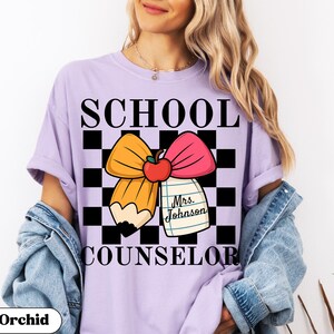 May include: A light purple t-shirt with the words "SCHOOL COUNSELOR" in black. The shirt features a graphic of a pencil, bow, and apple on a black and white checkered background. The text "Mrs. Johnson" is on a notepad.