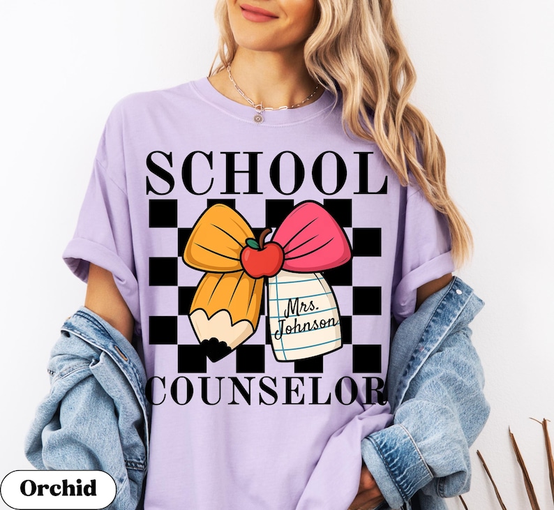 May include: A light purple t-shirt with the words "SCHOOL COUNSELOR" in black. The shirt features a graphic of a pencil, bow, and apple on a black and white checkered background. The text "Mrs. Johnson" is on a notepad.