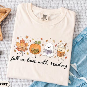 Peut inclure: Un t-shirt ivoire avec un motif repr&eacute;sentant une feuille d'&eacute;rable, une citrouille, un fant&ocirc;me et une tasse de caf&eacute;, chacun tenant un livre. Le texte "fall in love with reading" est imprim&eacute; sous les personnages, entour&eacute; de feuilles d'automne.