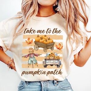 May include: A cream-colored t-shirt featuring a graphic print with the text "take me to the pumpkin patch." The design includes a wooden wagon filled with orange pumpkins, a scarecrow in plaid clothing, a blue pickup truck, and autumn leaves.