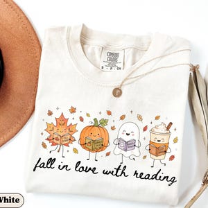 Peut inclure: Un t-shirt blanc &agrave; manches courtes avec une impression graphique repr&eacute;sentant une feuille d'&eacute;rable, une citrouille, un fant&ocirc;me et une tasse de caf&eacute; souriants, tenant chacun un livre. Le texte indique fall in love with reading.