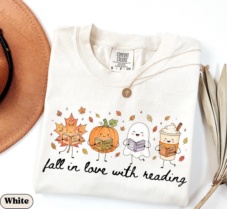 Peut inclure: Un t-shirt blanc &agrave; manches courtes avec une impression graphique repr&eacute;sentant une feuille d'&eacute;rable, une citrouille, un fant&ocirc;me et une tasse de caf&eacute; souriants, tenant chacun un livre. Le texte indique fall in love with reading.