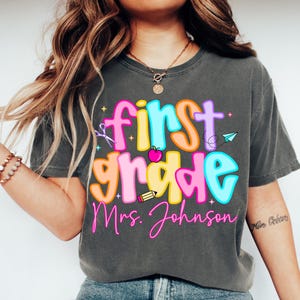May include: Dark grey t-shirt with a colorful "First Grade Mrs. Johnson" graphic. The design includes a pencil, apple, scissors, and paper airplane. The text is in bright pink, orange, yellow, and blue.