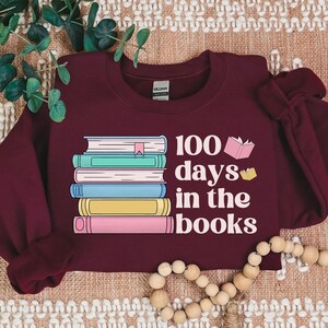 Pu&ograve; includere: Felpa color borgogna con una pila di libri colorati e il testo "100 giorni tra i libri".