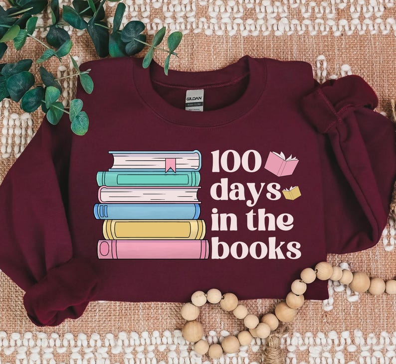 Pu&ograve; includere: Felpa color borgogna con una pila di libri colorati e il testo "100 giorni tra i libri".
