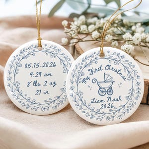 May include: Two round white ceramic ornaments with gold hanging strings. One features a blue floral wreath surrounding birth details: 05.15.2026, 9:29 am, 8 lbs 2 oz, 21 in. The other shows a blue baby carriage, My First Christmas, Liam Noah, 2026.