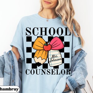 May include: Light blue t-shirt with the words "SCHOOL COUNSELOR" in black block letters. The shirt features a graphic of a yellow pencil and pink bow with a red apple in the center, set against a black and white checkered background. A notepad with the name "Mrs. Johnson" is also included.