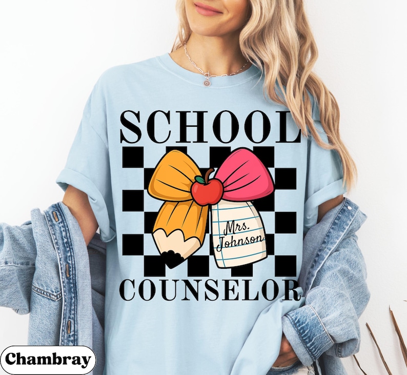 May include: Light blue t-shirt with the words "SCHOOL COUNSELOR" in black block letters. The shirt features a graphic of a yellow pencil and pink bow with a red apple in the center, set against a black and white checkered background. A notepad with the name "Mrs. Johnson" is also included.