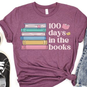 Pu&ograve; includere: Una maglietta viola con il testo "100 giorni tra i libri" e una pila di libri colorati con un segnalibro in cima.