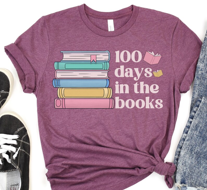 Pu&ograve; includere: Una maglietta viola con il testo "100 giorni tra i libri" e una pila di libri colorati con un segnalibro in cima.