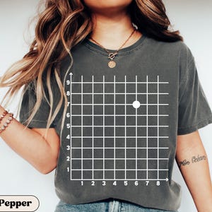 May include: Dark grey t-shirt featuring a white grid design with a single white dot. The shirt has a crew neck and short sleeves. The word "Pepper" is printed at the bottom of the shirt.