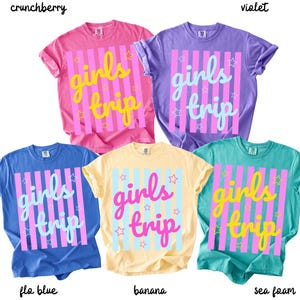 May include: A collection of five short-sleeved t-shirts in various colors: pink, purple, blue, yellow, and teal. Each shirt features the text "girls trip" in a contrasting color, with vertical stripes and star accents.