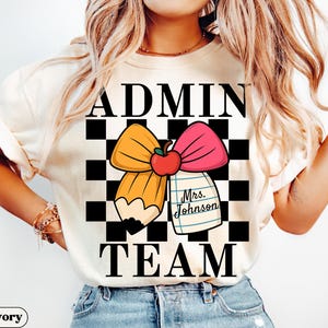 May include: Ivory-colored t-shirt with the words "ADMIN TEAM" in black block letters. A pencil and bow graphic with an apple and a notepad that says "Mrs. Johnson" is in the center. The background is a black and white checkered pattern.