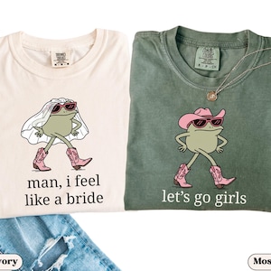 May include: Two t-shirts, one ivory and one moss green, featuring frog illustrations. The ivory shirt has a frog in a veil and cowboy boots with the text "man, I feel like a bride." The green shirt has a frog in a cowboy hat and boots with the text "let's go girls."