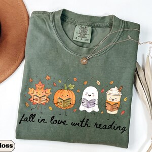 Peut inclure: Un t-shirt &agrave; col rond vert mousse avec un motif repr&eacute;sentant une feuille d'&eacute;rable, une citrouille, un fant&ocirc;me et une tasse de caf&eacute;, chacun tenant un livre. Le texte "fall in love with reading" est imprim&eacute; sous les personnages.