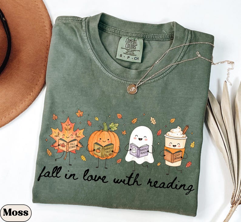 Peut inclure: Un t-shirt &agrave; col rond vert mousse avec un motif repr&eacute;sentant une feuille d'&eacute;rable, une citrouille, un fant&ocirc;me et une tasse de caf&eacute;, chacun tenant un livre. Le texte "fall in love with reading" est imprim&eacute; sous les personnages.