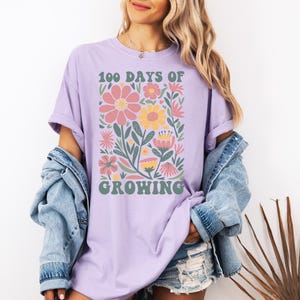 May include: A lavender t-shirt with a graphic design of colorful flowers and the text "100 Days of Growing" in a retro font.