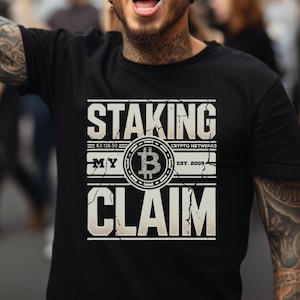Puede incluir: Un hombre con una camiseta negra con un diseño gráfico blanco que dice "Staking My Claim" con un símbolo de Bitcoin. El gráfico también incluye el texto "Crypto Networks Est. 2008" y "63,710,501".