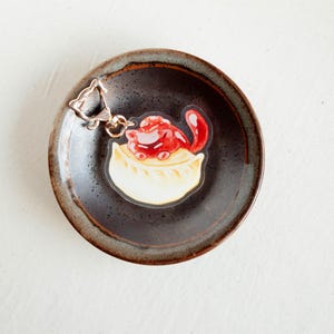 May include: A clear acrylic keychain featuring a cartoon dumpling with a red topping. The keychain is attached to a silver-colored clasp and rests on a dark brown ceramic plate with a textured rim.