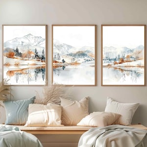 May include: Three watercolour paintings of a mountain lake scene with snow-capped mountains, trees, and reflections in the water. The paintings are framed in wood and hung on a white wall.