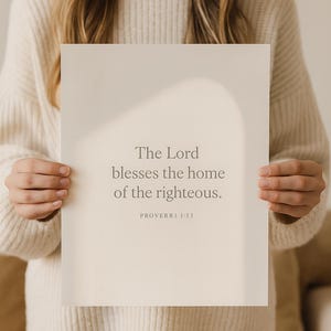 May include: A beige print with the text "The Lord blesses the home of the righteous. PROVERBS 3:33." The print is held by a person wearing a cream-colored sweater.
